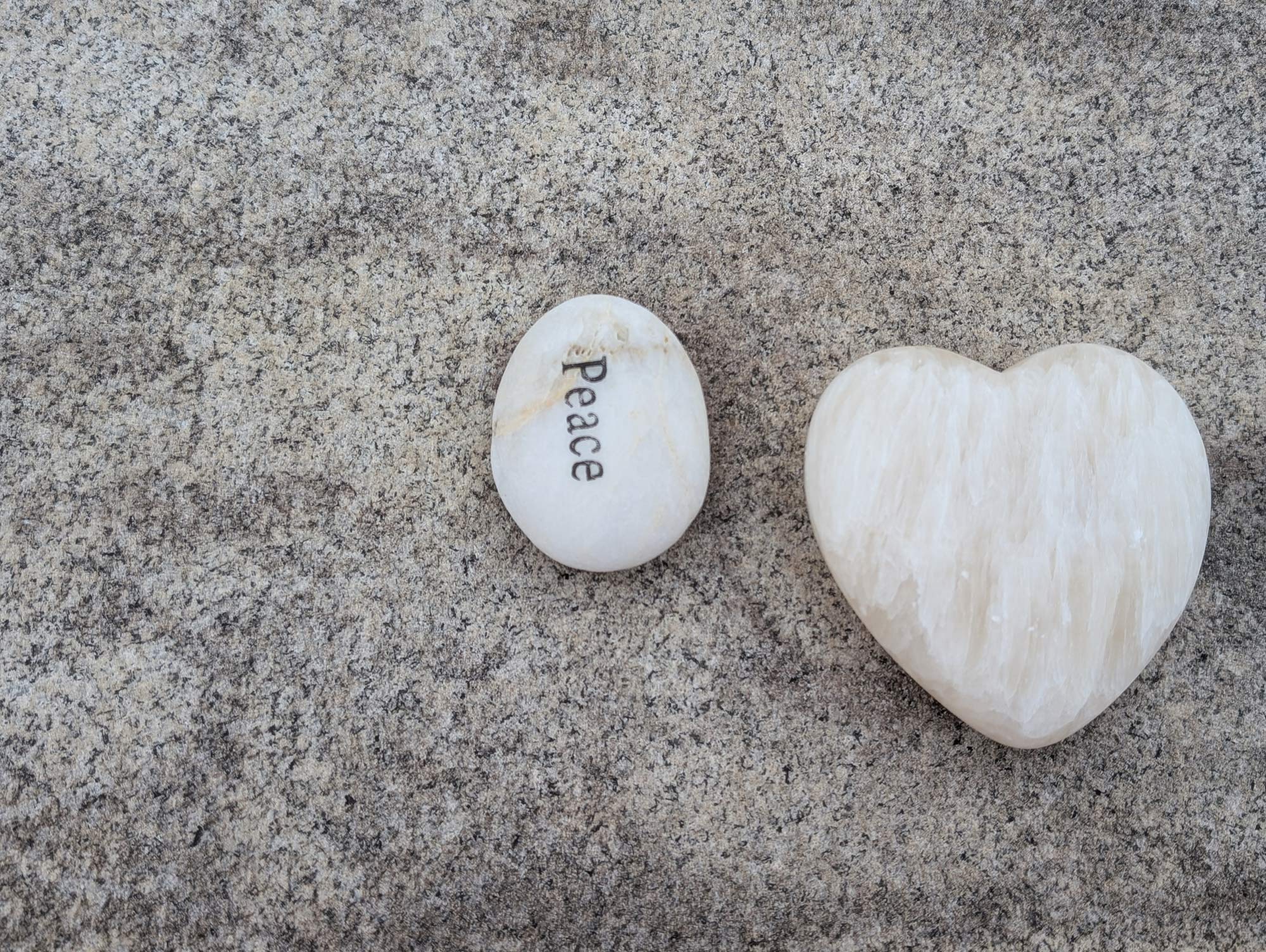 A white heart shaped stone, sitting beside another oval shaped stone with the word "PEACE" engraved on it, on the top of a grey granite tombstone