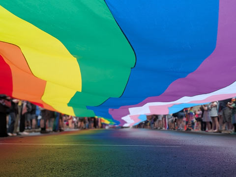 The underside of the train of a gown, colored like a Pride flag, the length and width of a city block, floating about 2-3 feet off of the ground