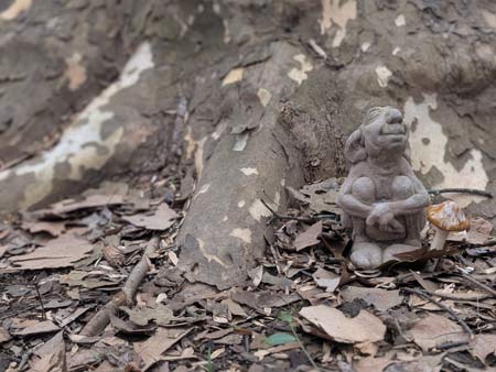 A cement figure of a gnome sniffing the air while sitting next to a ceramic mushroom at the base of a Sicamore tree