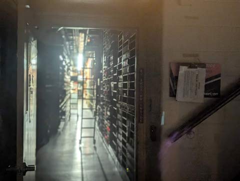 Photo through a door window of a doorway opening to a long corridor lined on each side with tall banks of telecommunications equipment, and two ladders that reach to tracks on the ceiling and have wheels at the bottom on the floor